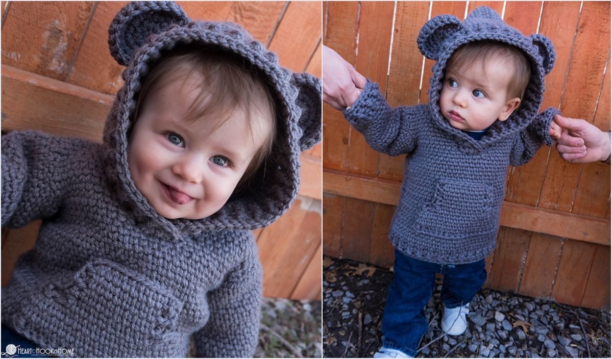 A baby wearing a gray knitted sweater with bear ears, reminiscent of an amigurumi stitch, is held by an adult while standing outdoors by a wooden fence.
