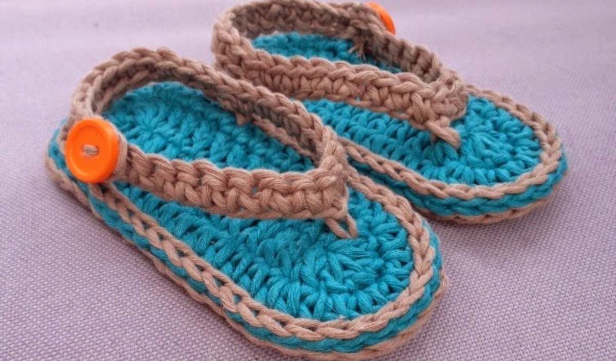 Discover adorable crocheted blue and beige baby flip-flop sandals with charming orange button accents, perched elegantly on a gray surface. Perfect for your little one's tiny toes!