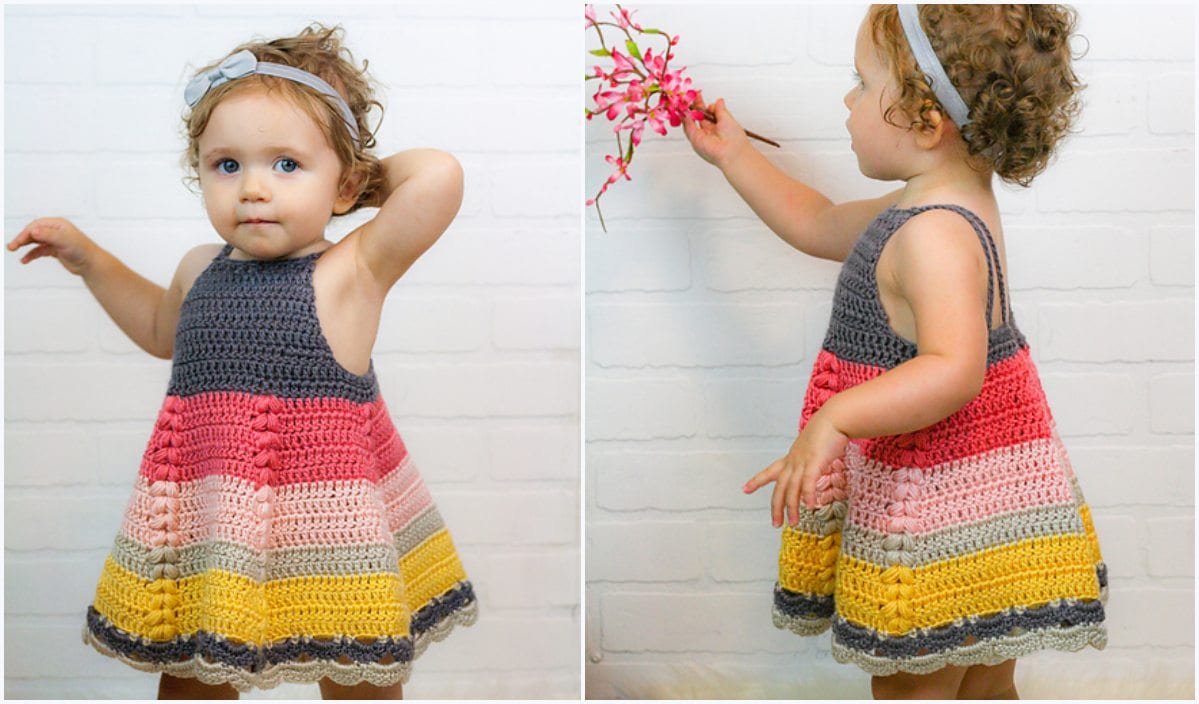 A toddler in a vibrant, puff stitch crocheted dress leans against a white brick wall, clutching a pink flower branch.