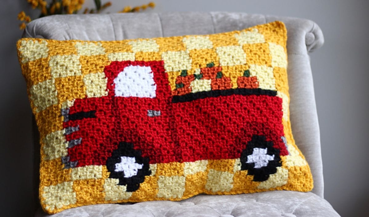 A decorative pillow featuring a charming red truck carrying pumpkins, expertly crocheted on a yellow and white checkered background, perfect for adding seasonal flair. Ideal for crochet enthusiasts looking to explore C2C pillow techniques with free crochet patterns available online.